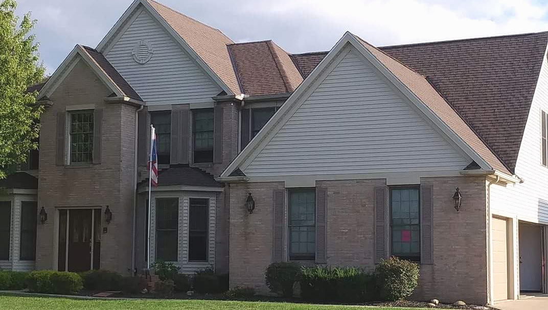 Two-story brick house with brown roof and beige siding, surrounded by green lawn and trees.