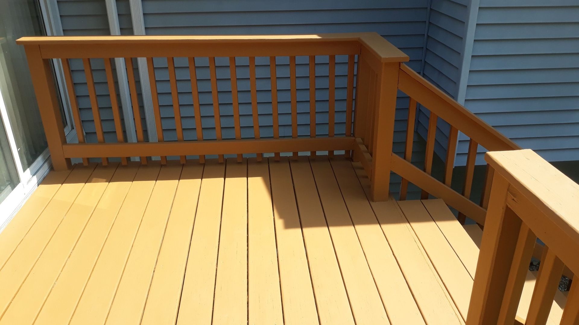 Wooden deck with railings, tan flooring, and brown railings, next to a blue siding wall.