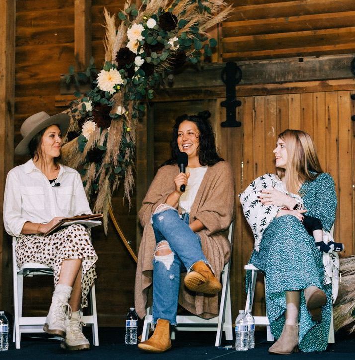 Three women are sitting in chairs talking to each other in front of a wooden wall.