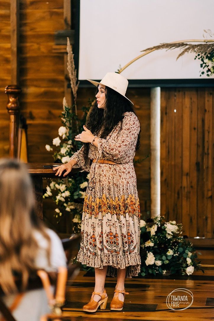 A woman in a dress and hat is standing in front of a crowd holding a microphone.