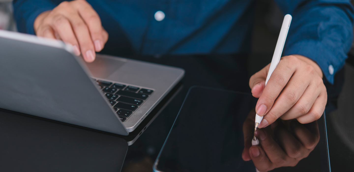 Person using a laptop and a tablet with a stylus on a dark surface.