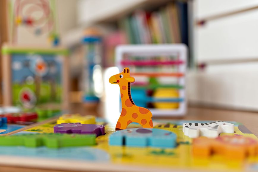 Wooden giraffe toy on a colorful play surface with other animal shapes, blurred background.
