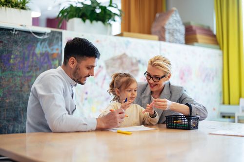 A man, child, and woman sit at a table drawing; brightly colored classroom.