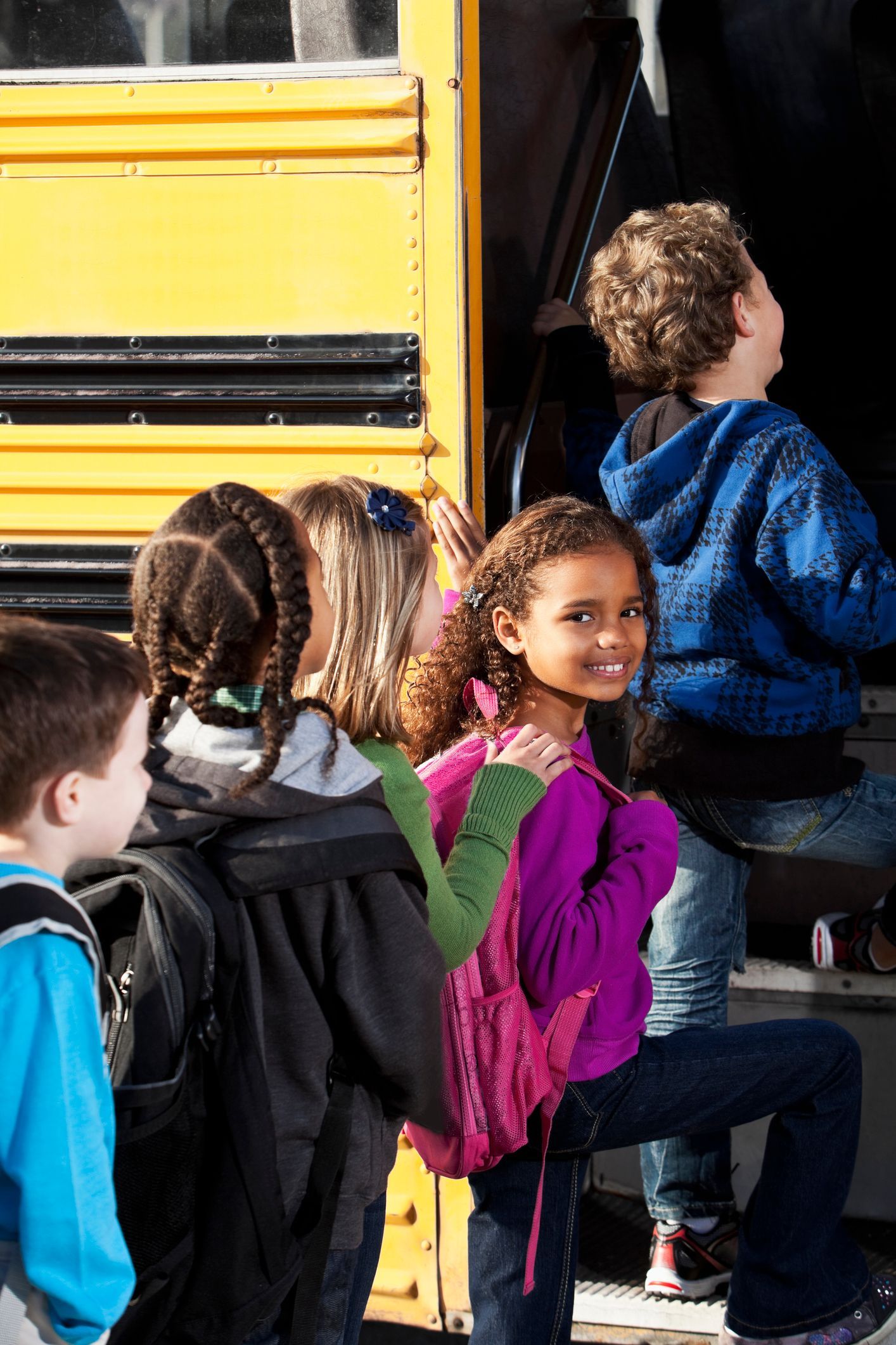 Children boarding a yellow school bus, one smiling at the camera, carrying backpacks.