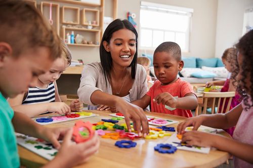 Teacher guides young students at a table sorting colorful shapes in a bright classroom.