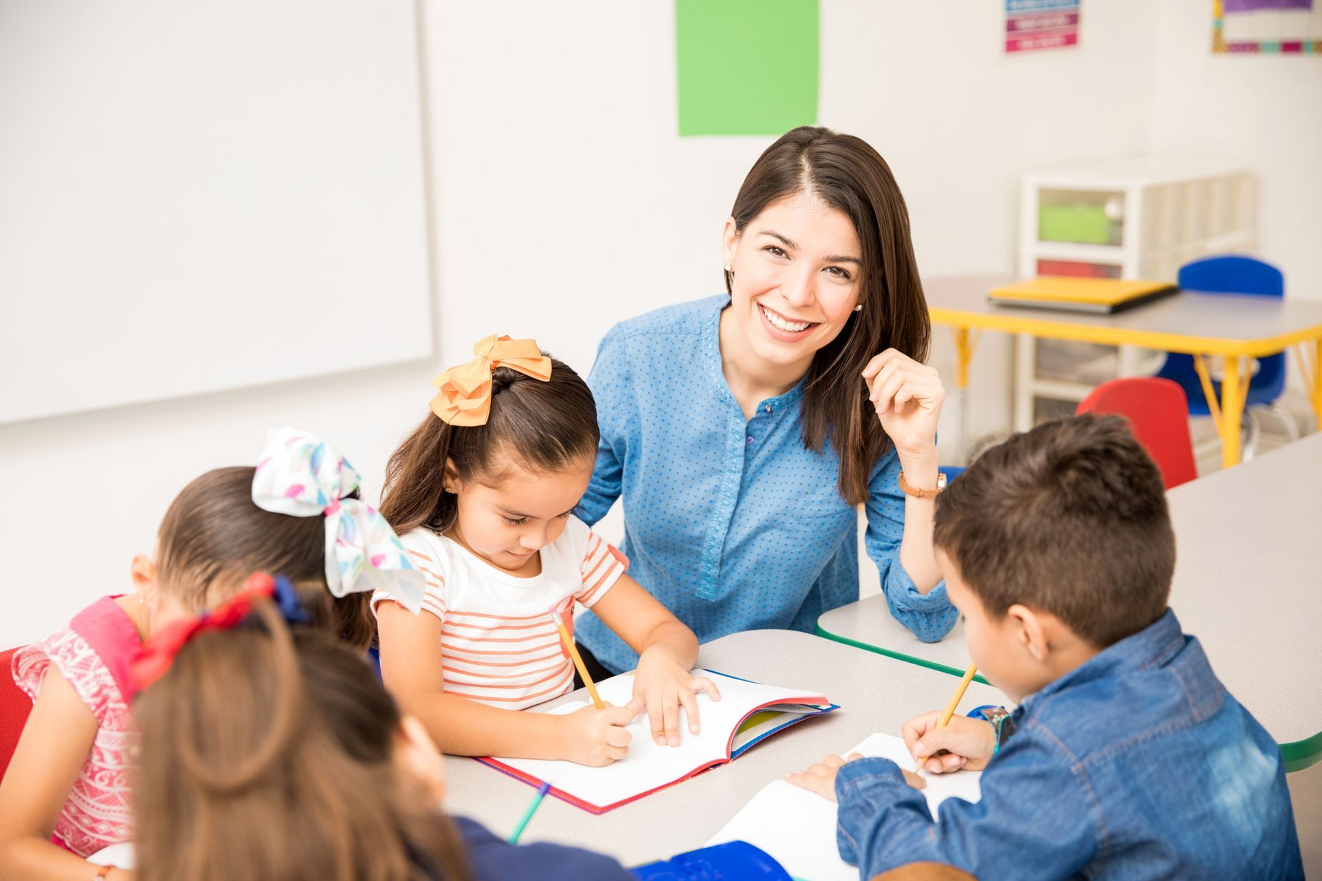 Teacher smiles while assisting young children writing in a classroom setting.