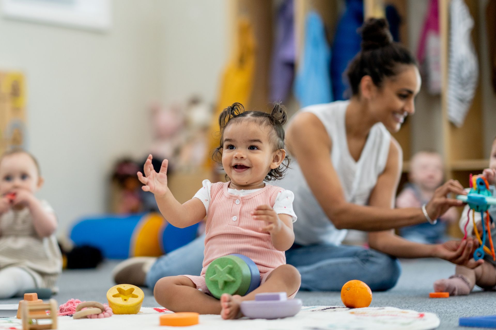 Happy toddler playing with colorful toys in a daycare, smiling at a teacher.