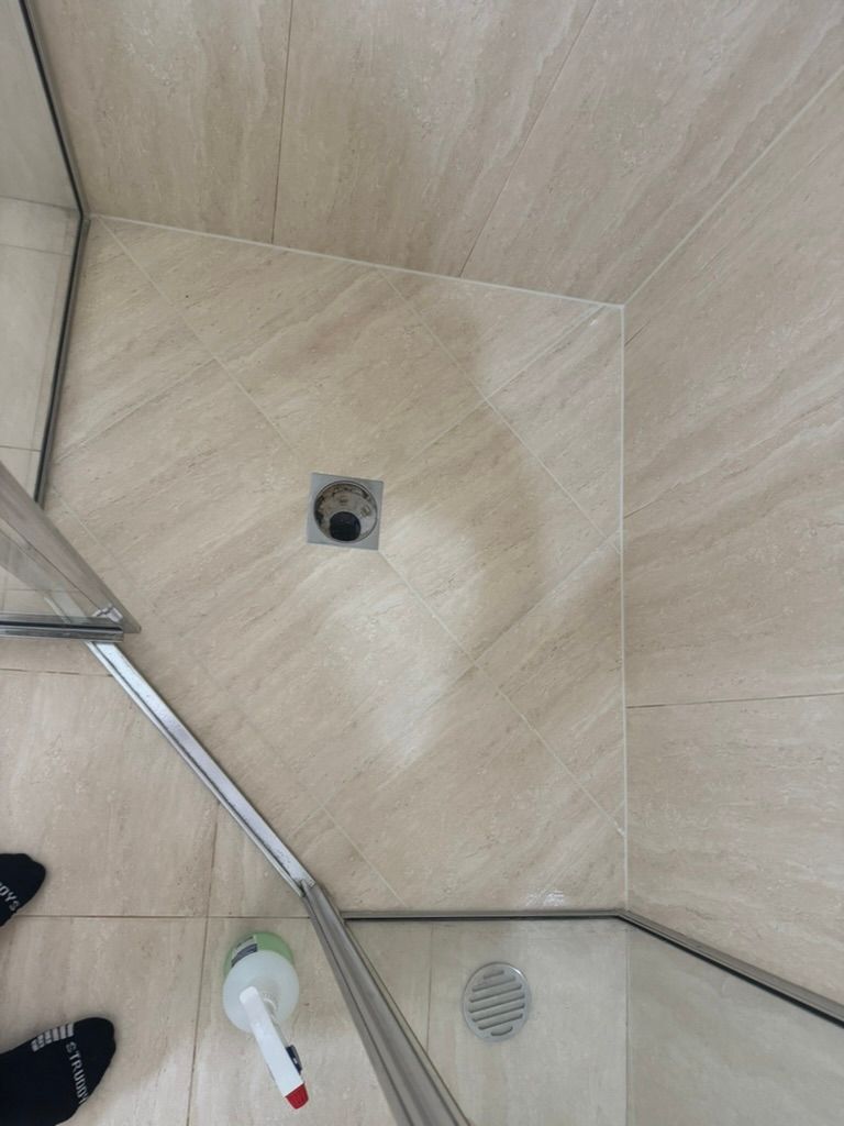 Shower floor with a drain and light beige tiles. A glass shower door is in the corner— 1 Time Waterproofing in East Ballina, NSW