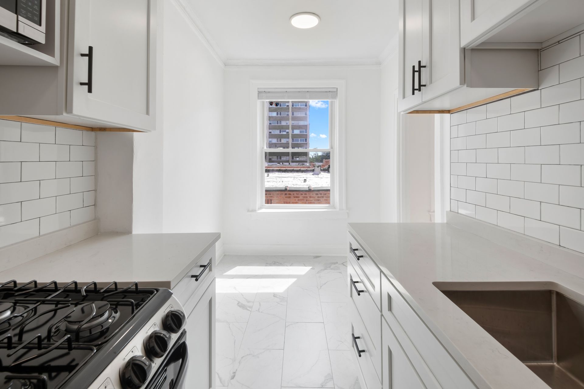 Narrow white kitchen with window. White cabinets, subway tile backsplash, gas stove, stainless steel sink.