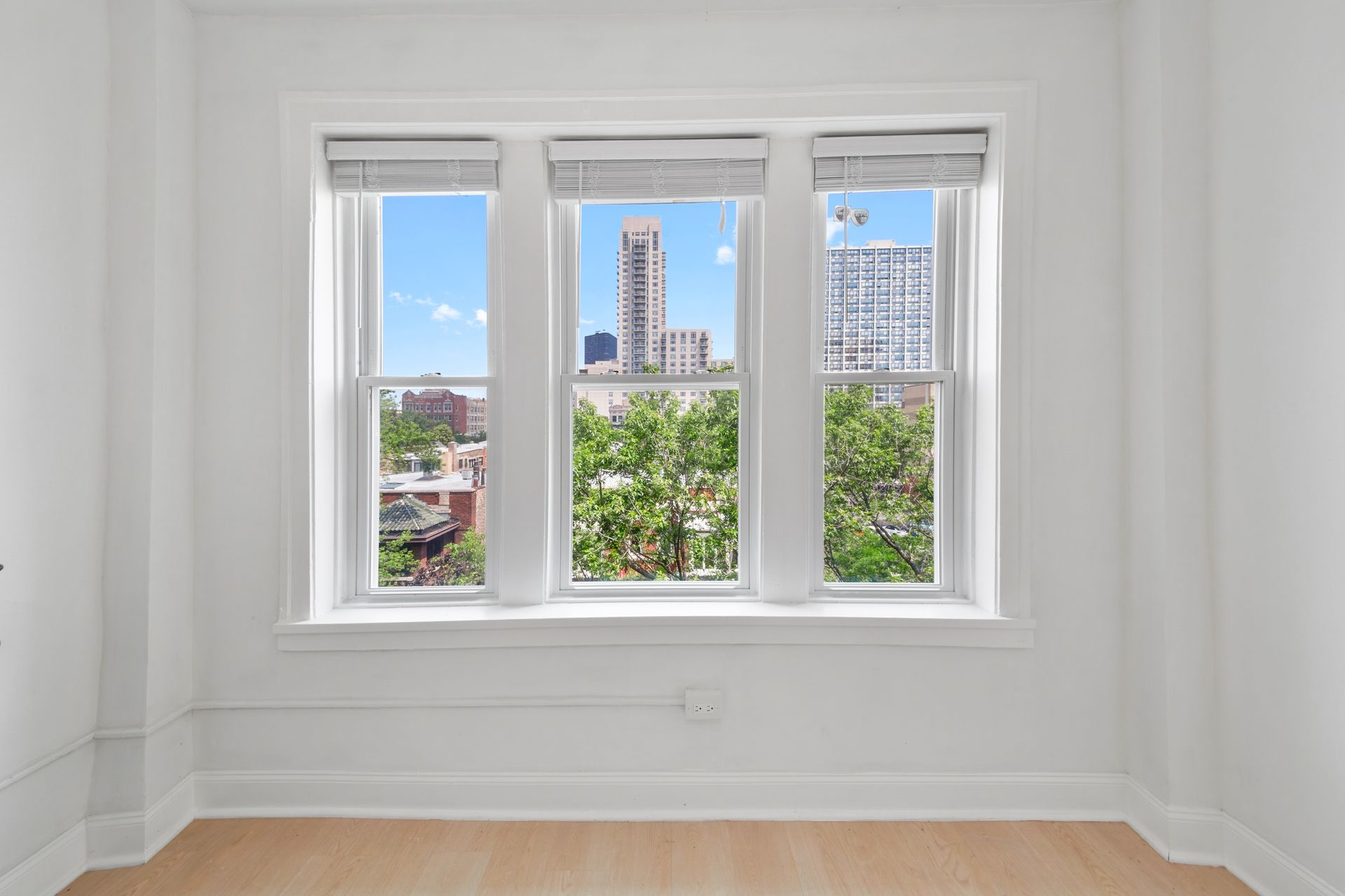 Bright room with a triple window overlooking a cityscape with trees and buildings. White walls, blinds, and wood floor.