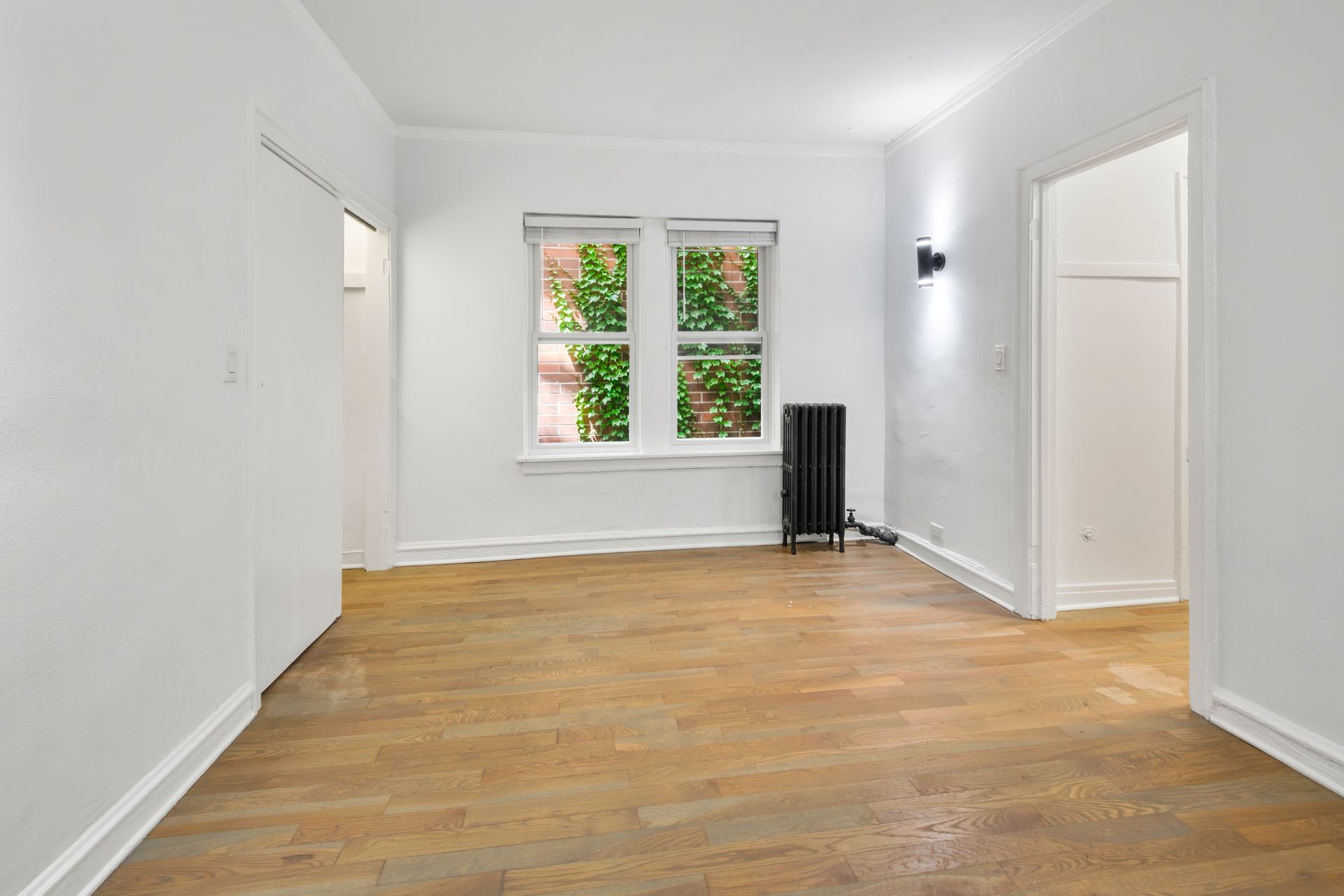 Empty room with wooden floor, white walls, window with greenery view, and black radiator.