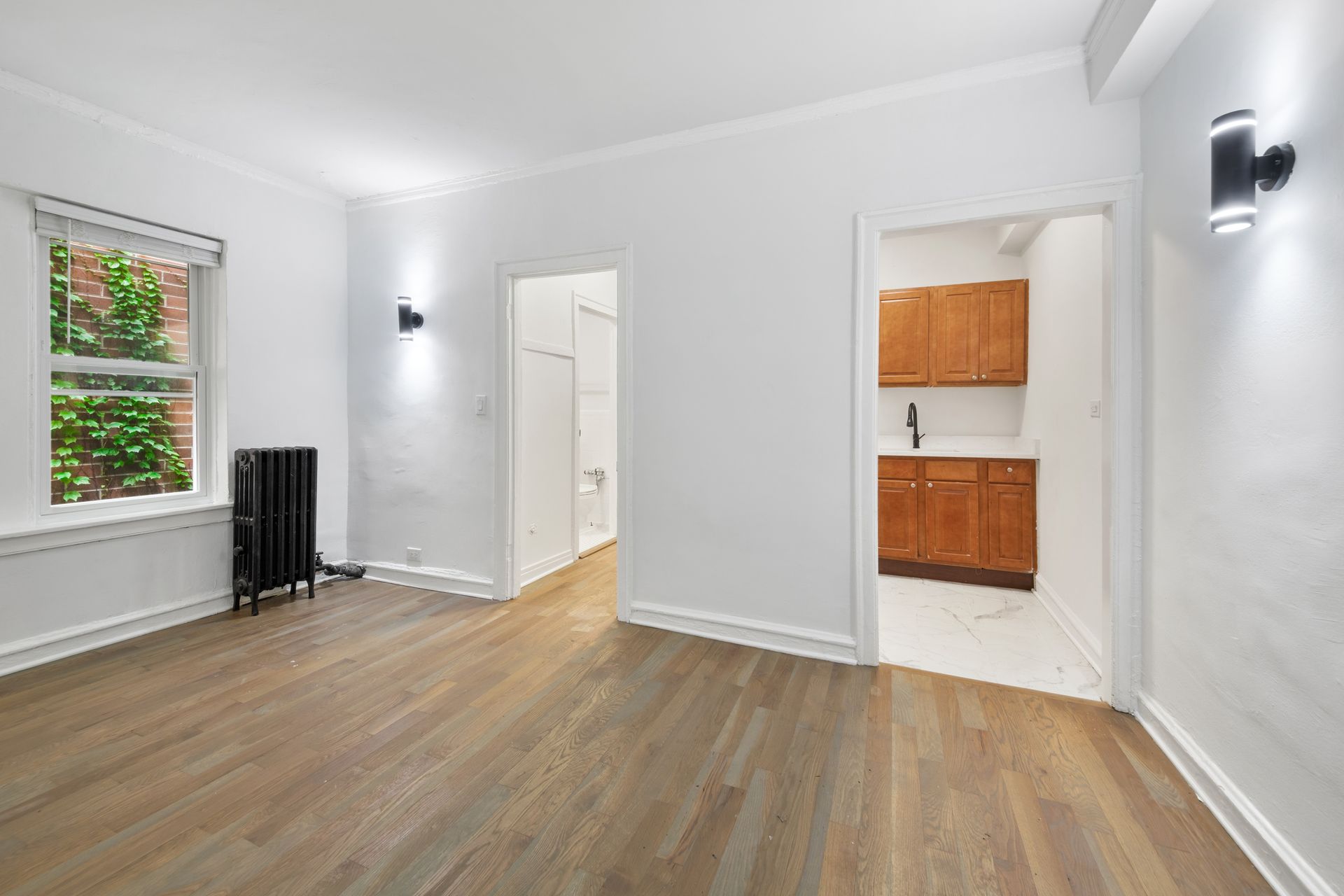 Empty room with hardwood floors, white walls, and a doorway to a kitchen with brown cabinets.
