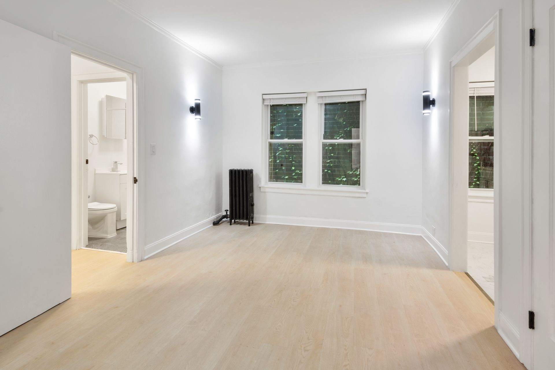 Empty white room with wood floor, window, radiator, and open doorways.