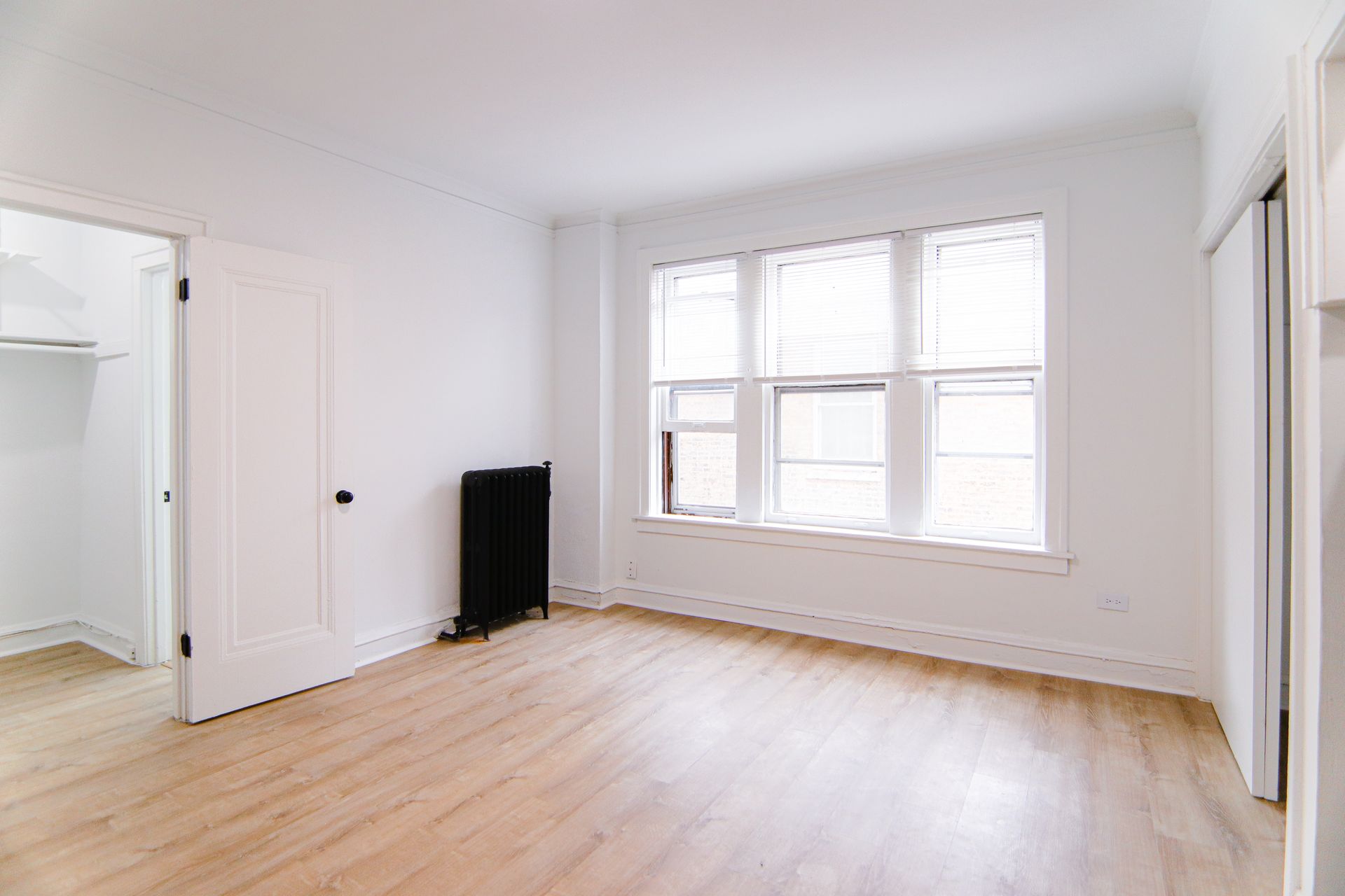 Empty room with light wood floors, large window with blinds, white walls, and a black radiator.