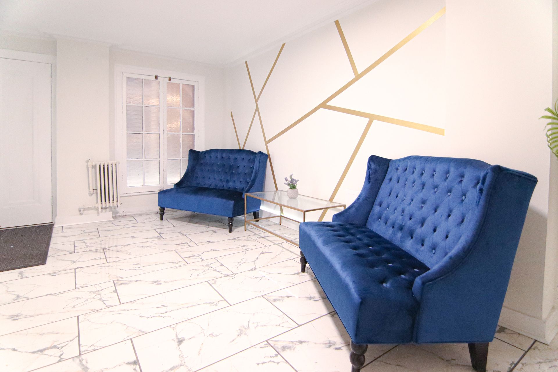 Two blue velvet sofas in a white room with gold geometric wall decor. Marble floor.