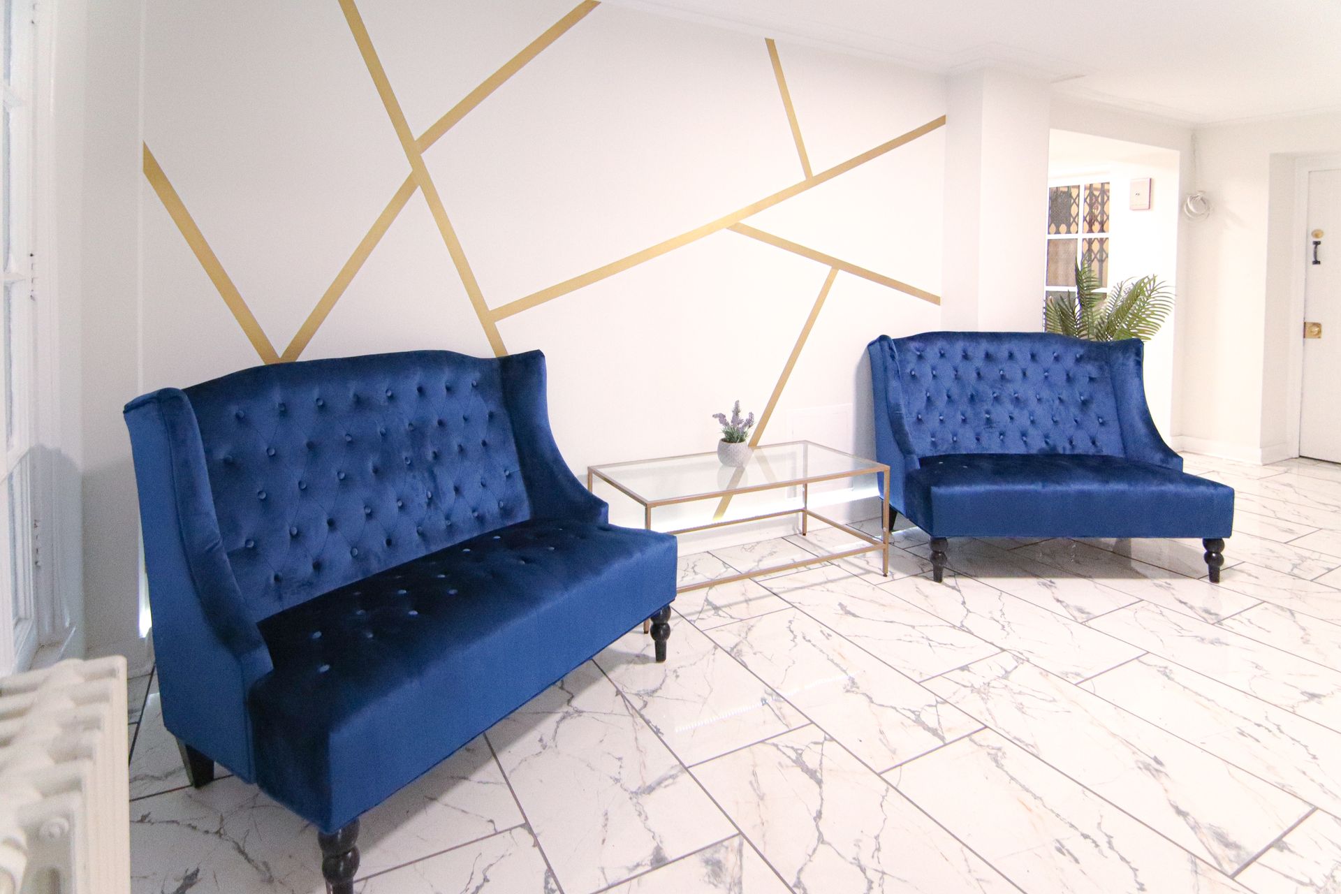 Two blue velvet sofas face each other in a bright room with white marble floors and gold geometric wall art.