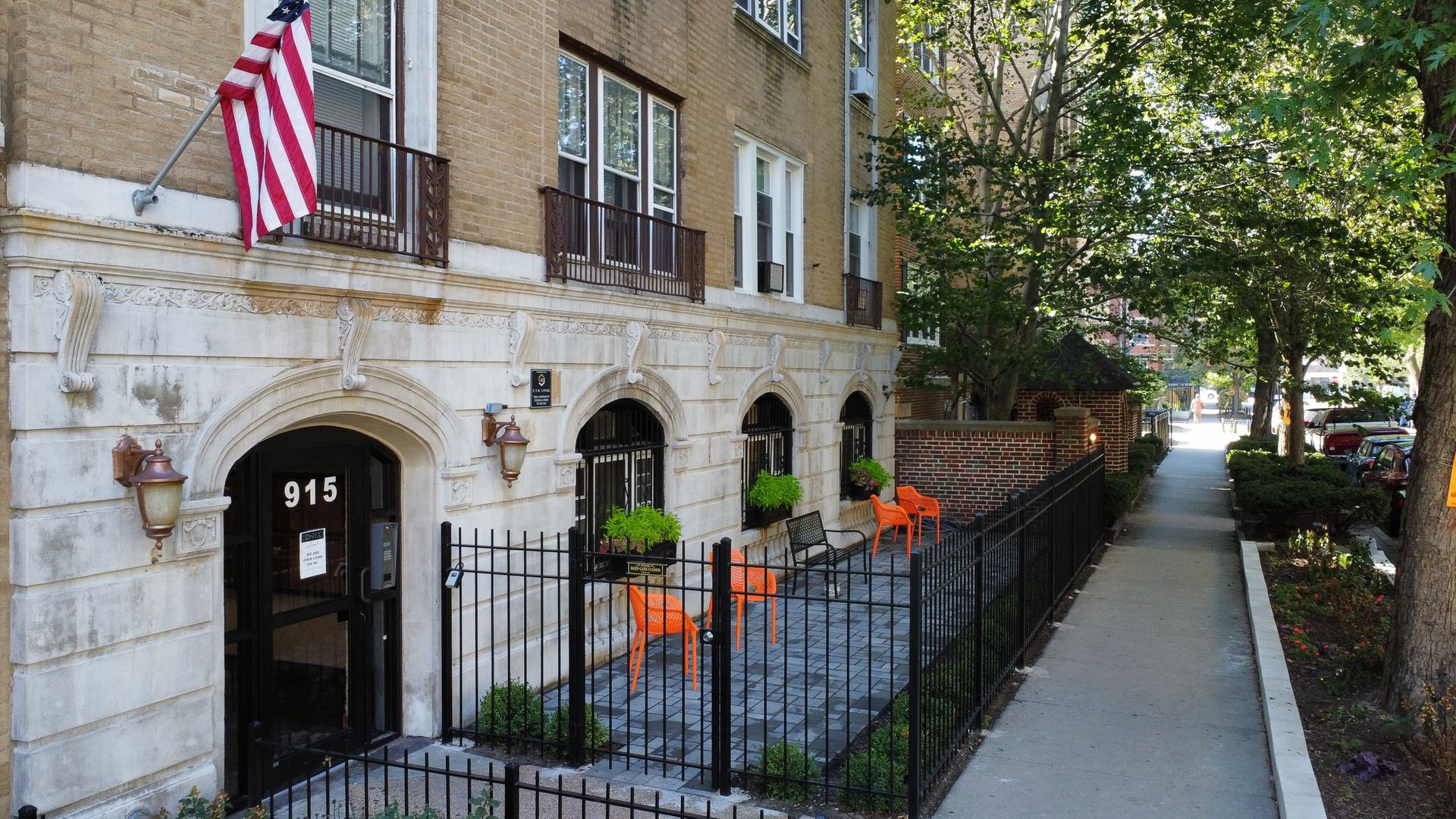 Apartment building entrance with black fence, orange chairs, and American flag.