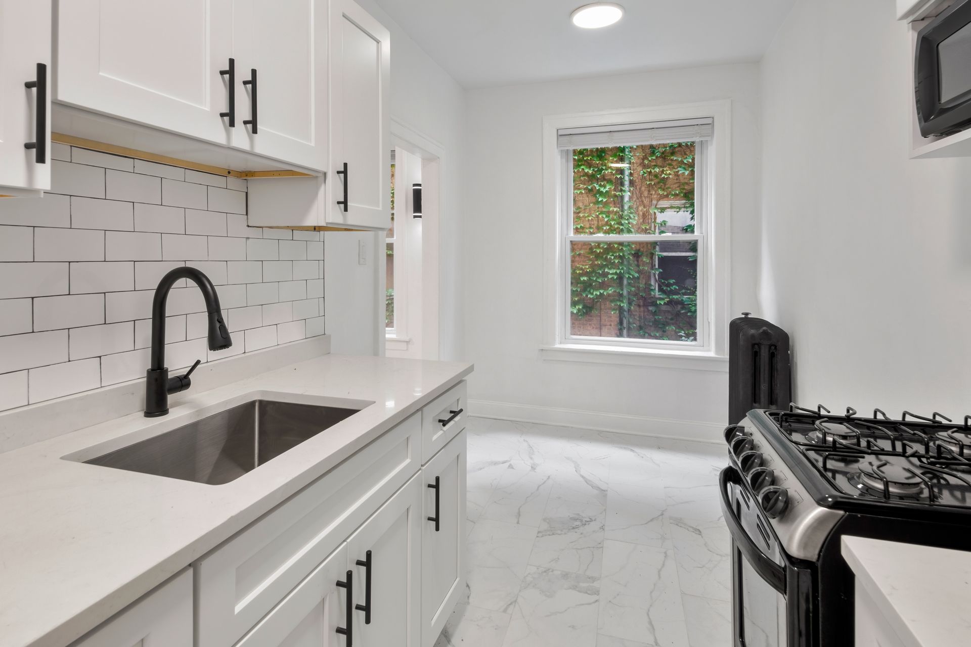 Bright white kitchen with sink, cabinets, and stove, with a window showing outdoor greenery.