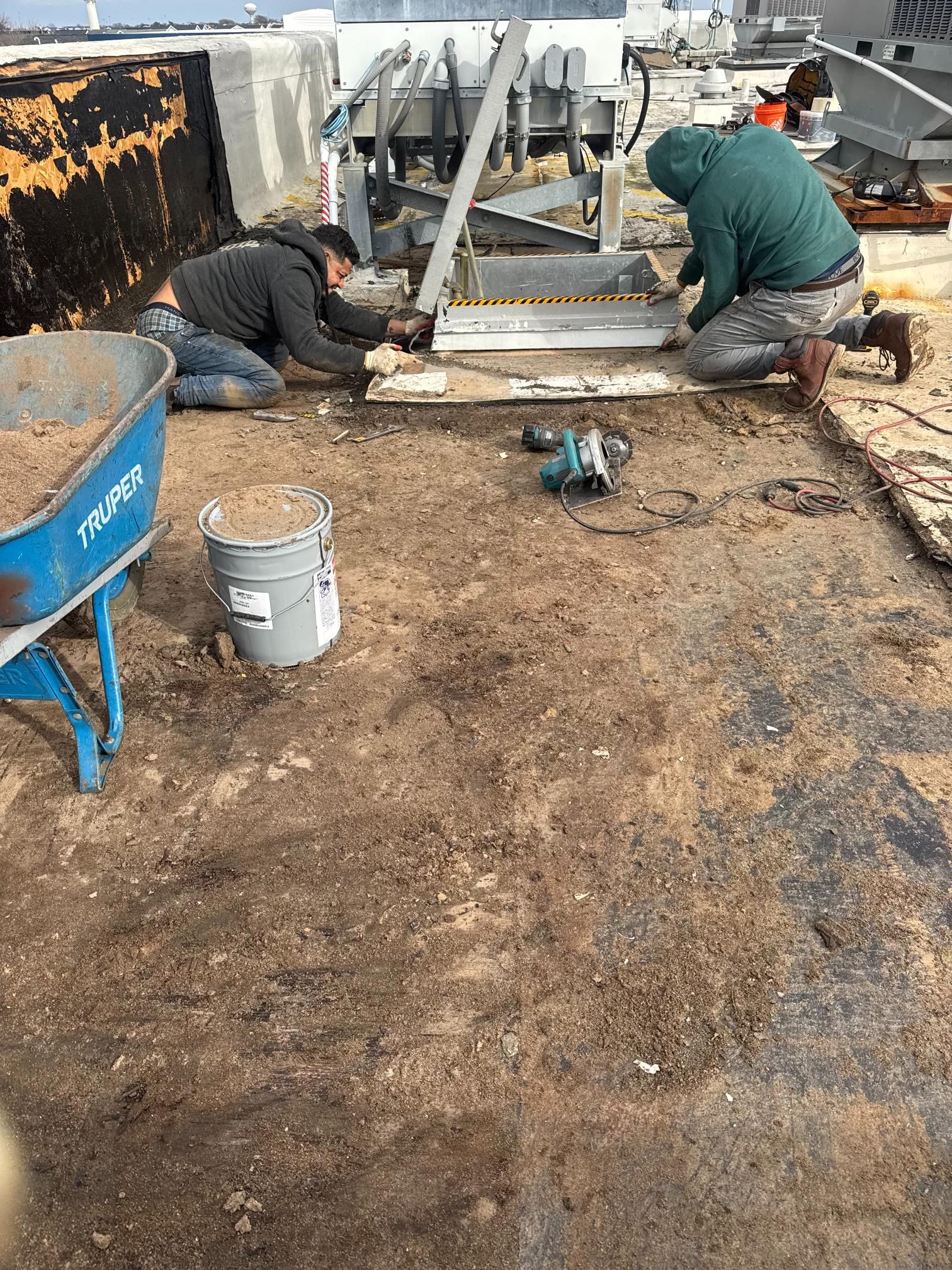 Two workers installing on a rooftop. One kneels, the other bends. Tools and a wheelbarrow nearby.