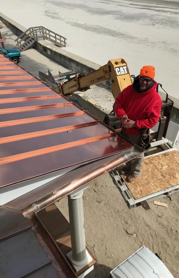 Man in orange hat and red jacket works on a copper roof. An excavator is in the background.