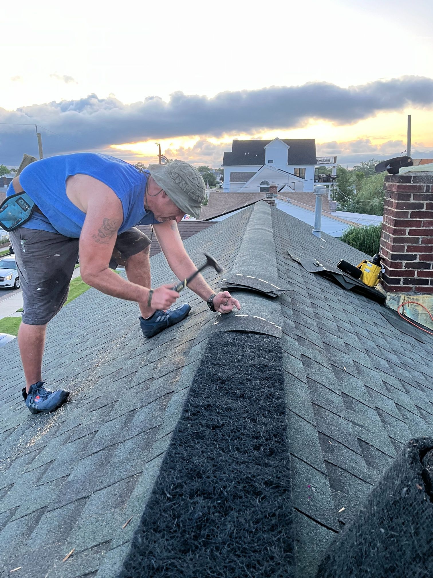 Man on roof, hammering shingles near chimney, with cloudy sky in the background.