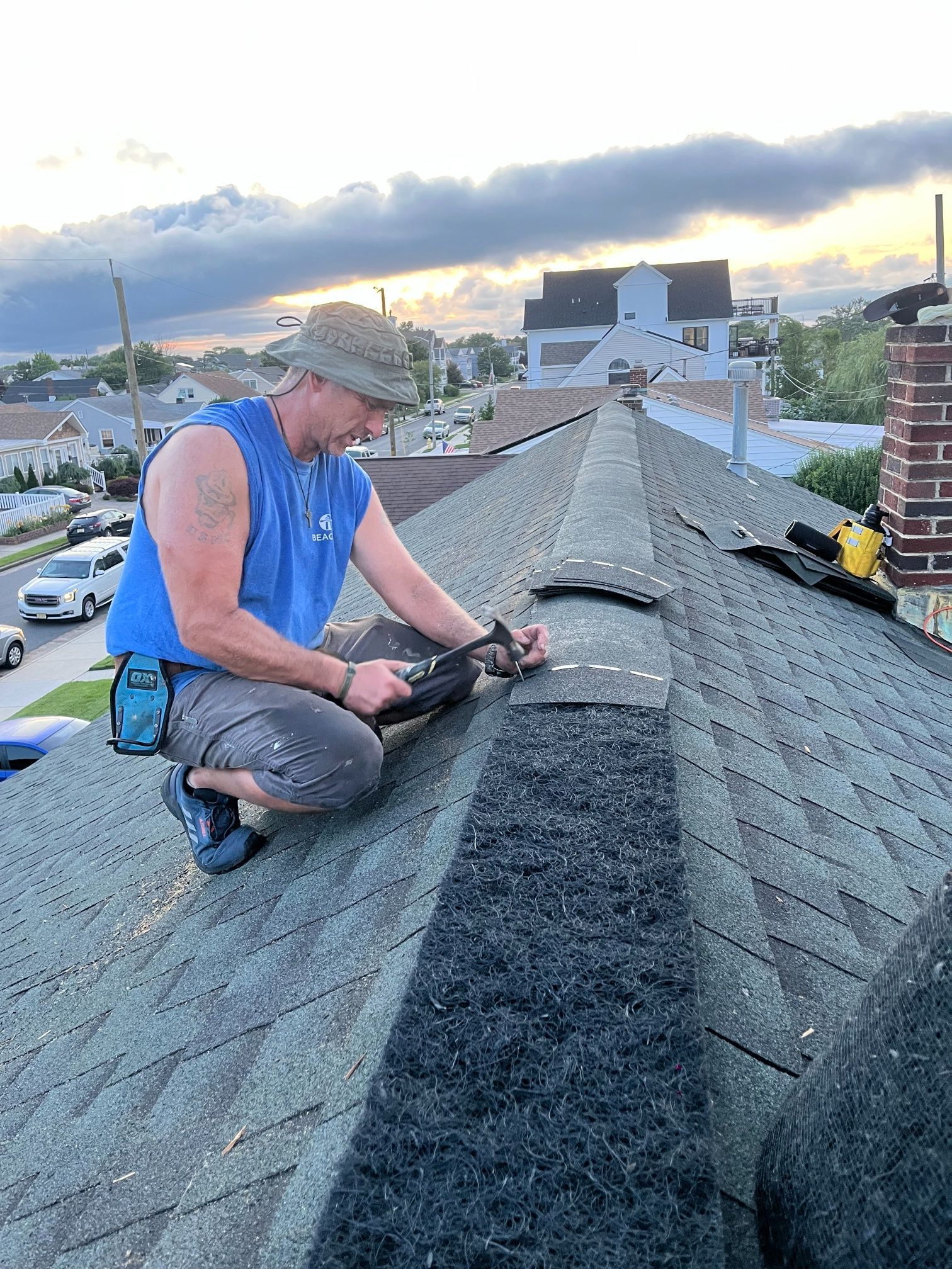 Roofer in a blue shirt and hat, works on a gray shingle roof, in front of houses at dusk.