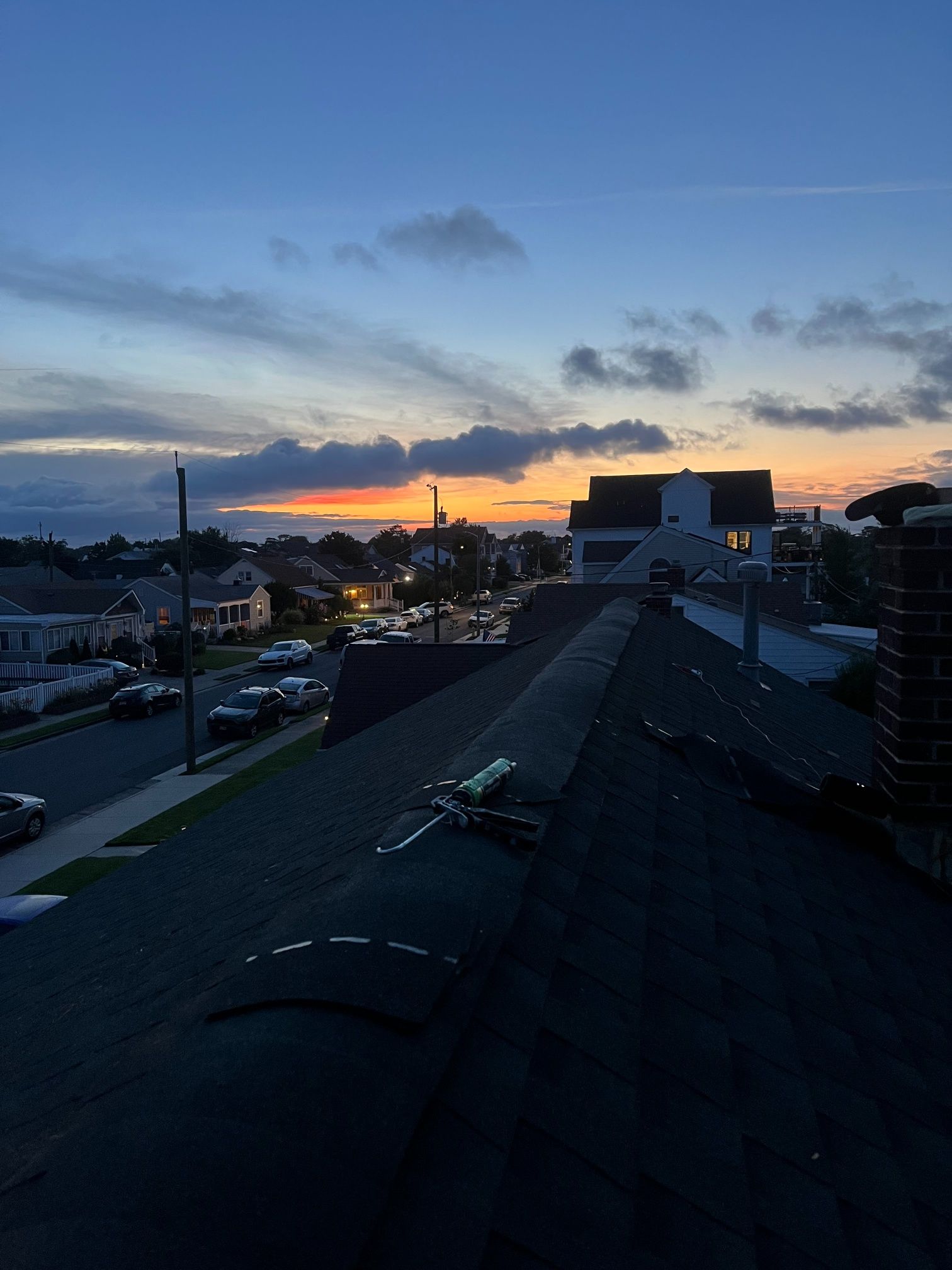 Rooftop view at dusk: street with cars, houses under orange and blue sky.