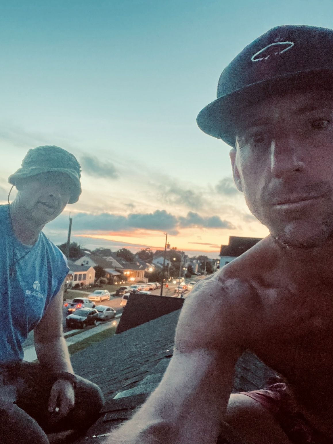 Two men on a rooftop at dusk. One shirtless, with a cap, taking a selfie; the other, wearing a hat, looking at the camera.