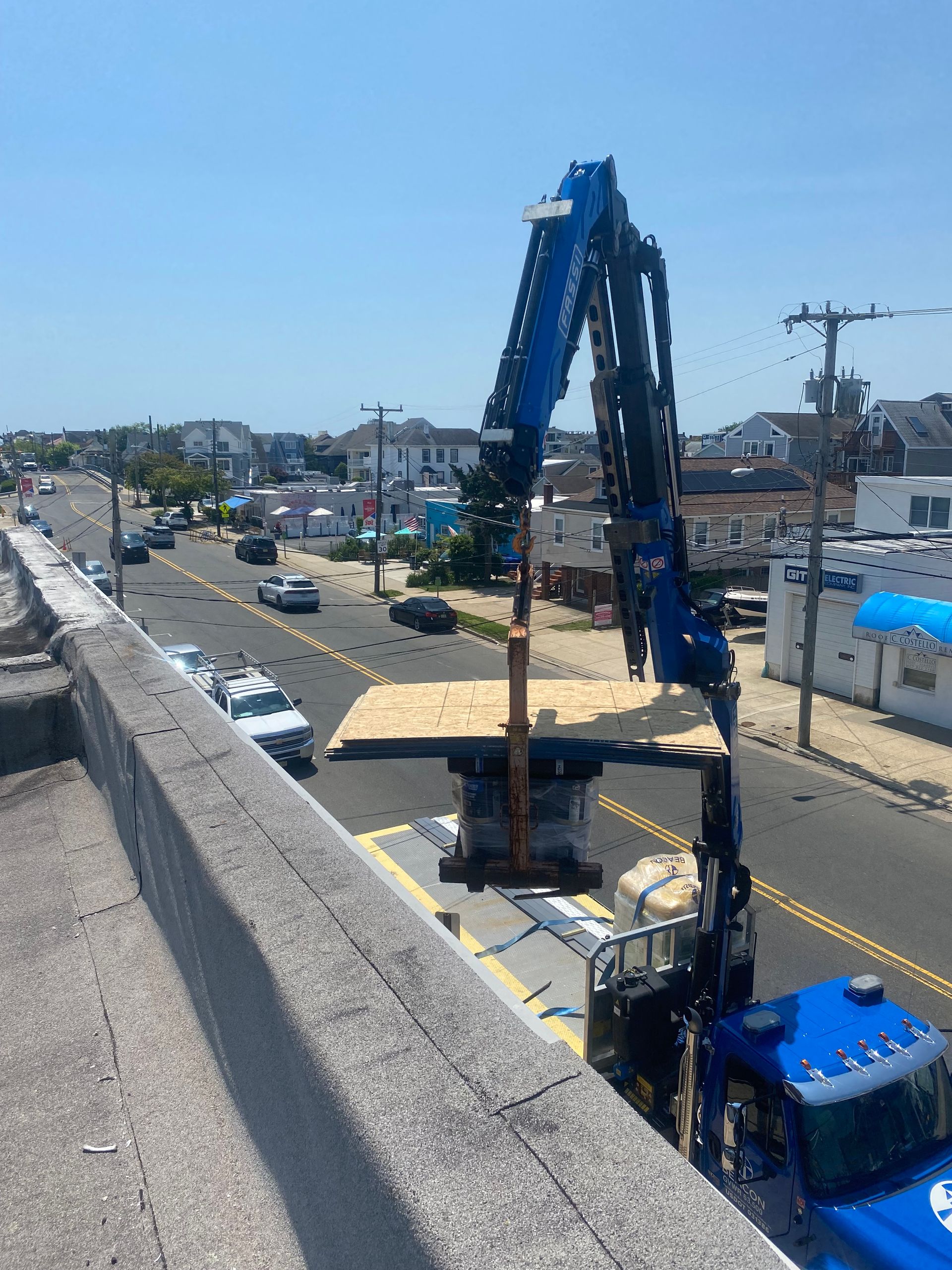 Crane lifting a wooden platform next to a road with cars and houses. Blue crane and truck, bright sunny day.