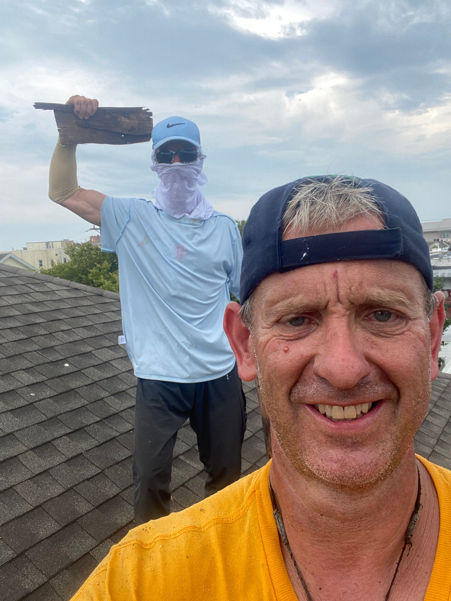 Two men on a rooftop. One holds a tool, other smiles at camera. Overcast sky.