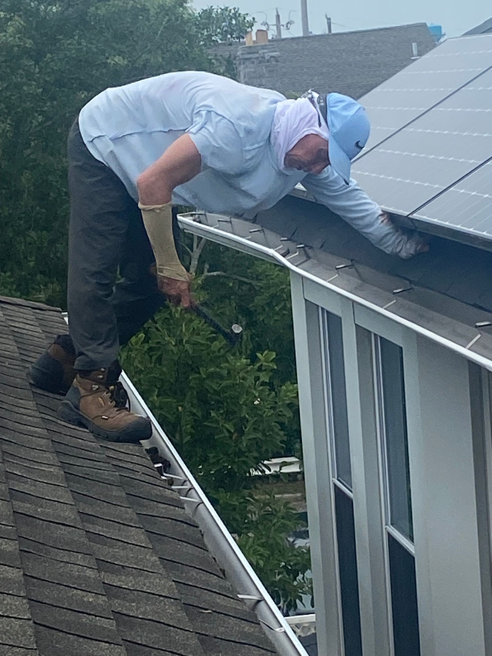 A man cleaning a gutter on a roof wearing a light blue shirt, cap, and gloves.