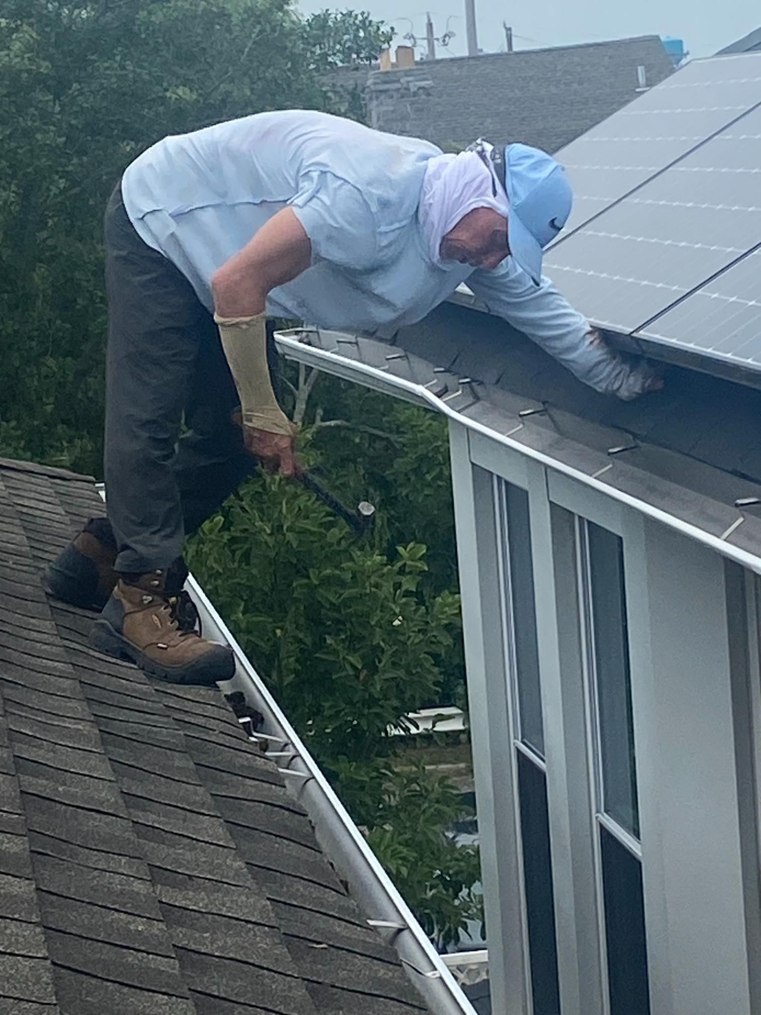A person cleans a gutter on a rooftop with solar panels. They wear a light blue shirt, cap, and gloves.