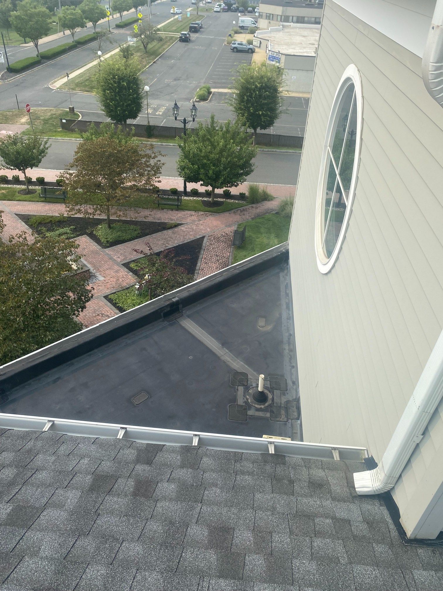View from a rooftop: asphalt shingles, flat black roof, round window, green trees, brick walkway, parking lot.