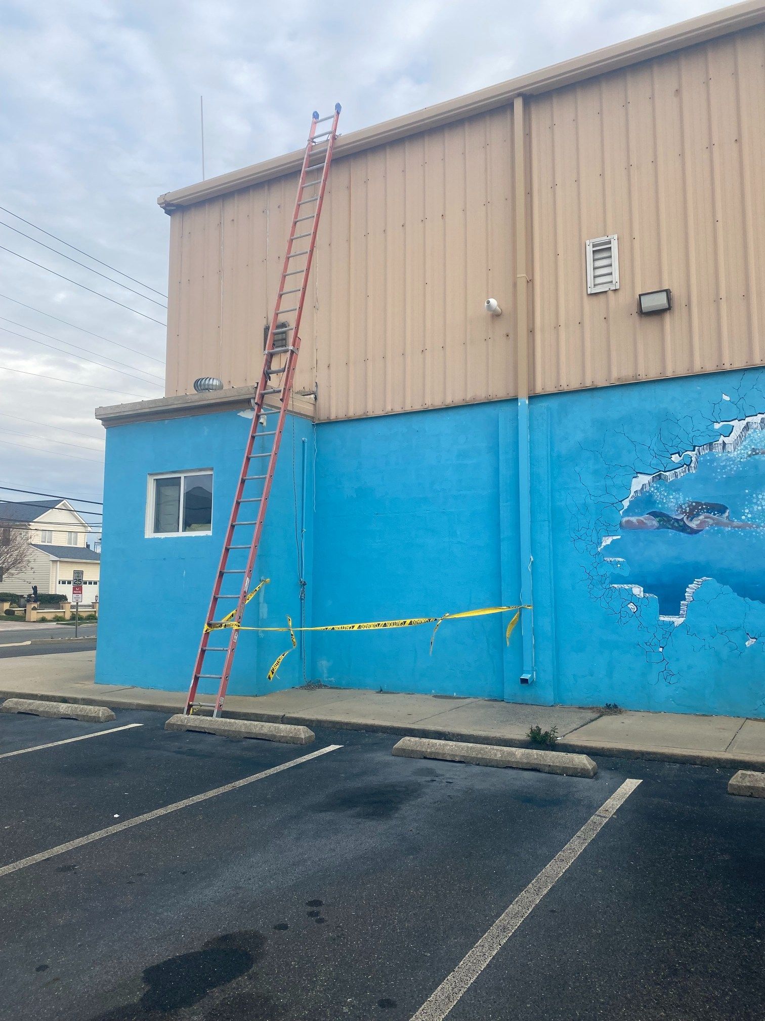 Tall ladder leaning against a building with blue and tan walls, yellow caution tape on the ground.