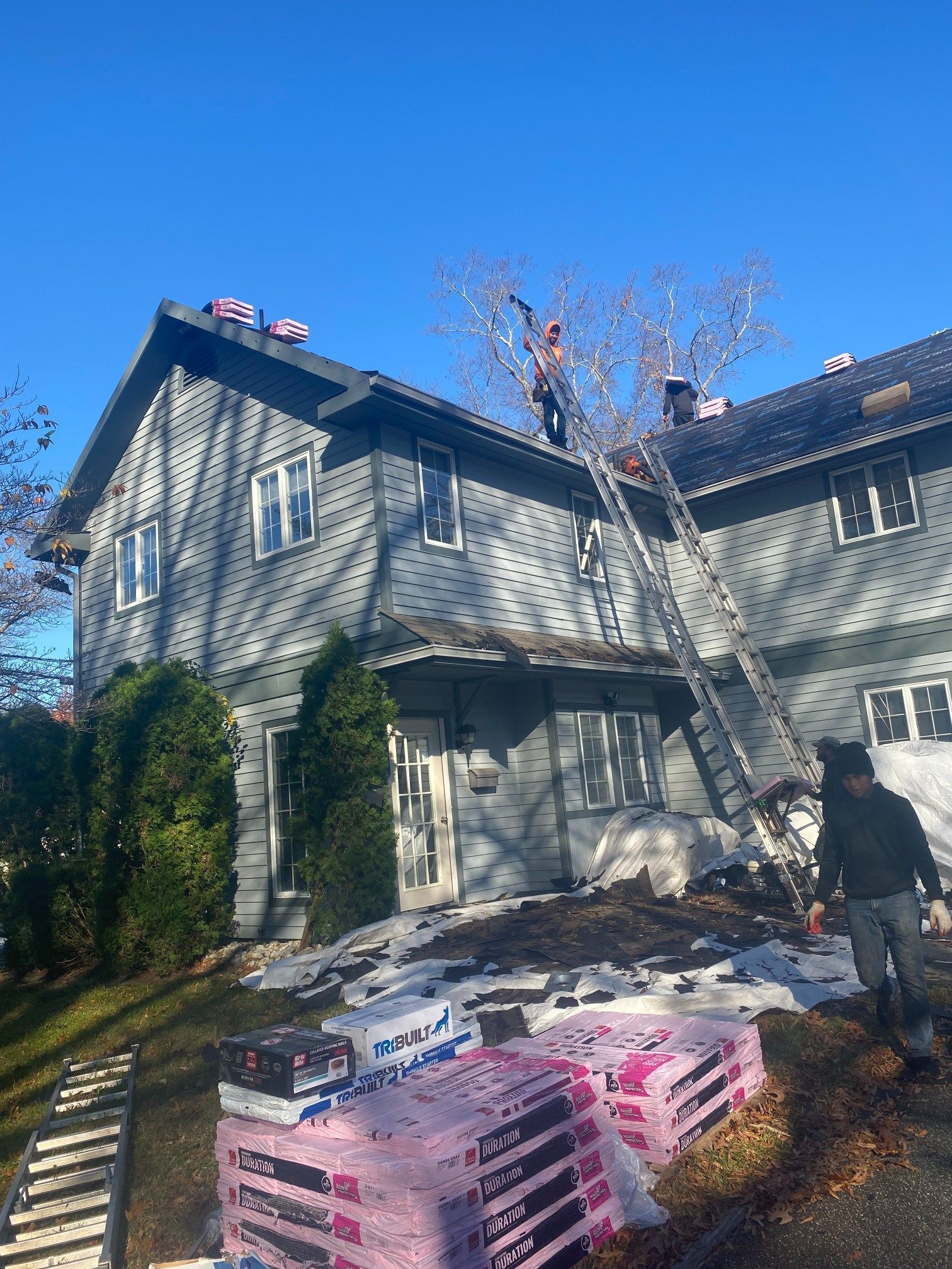 People on a roof with a ladder, installing shingles on a gray house under a blue sky.