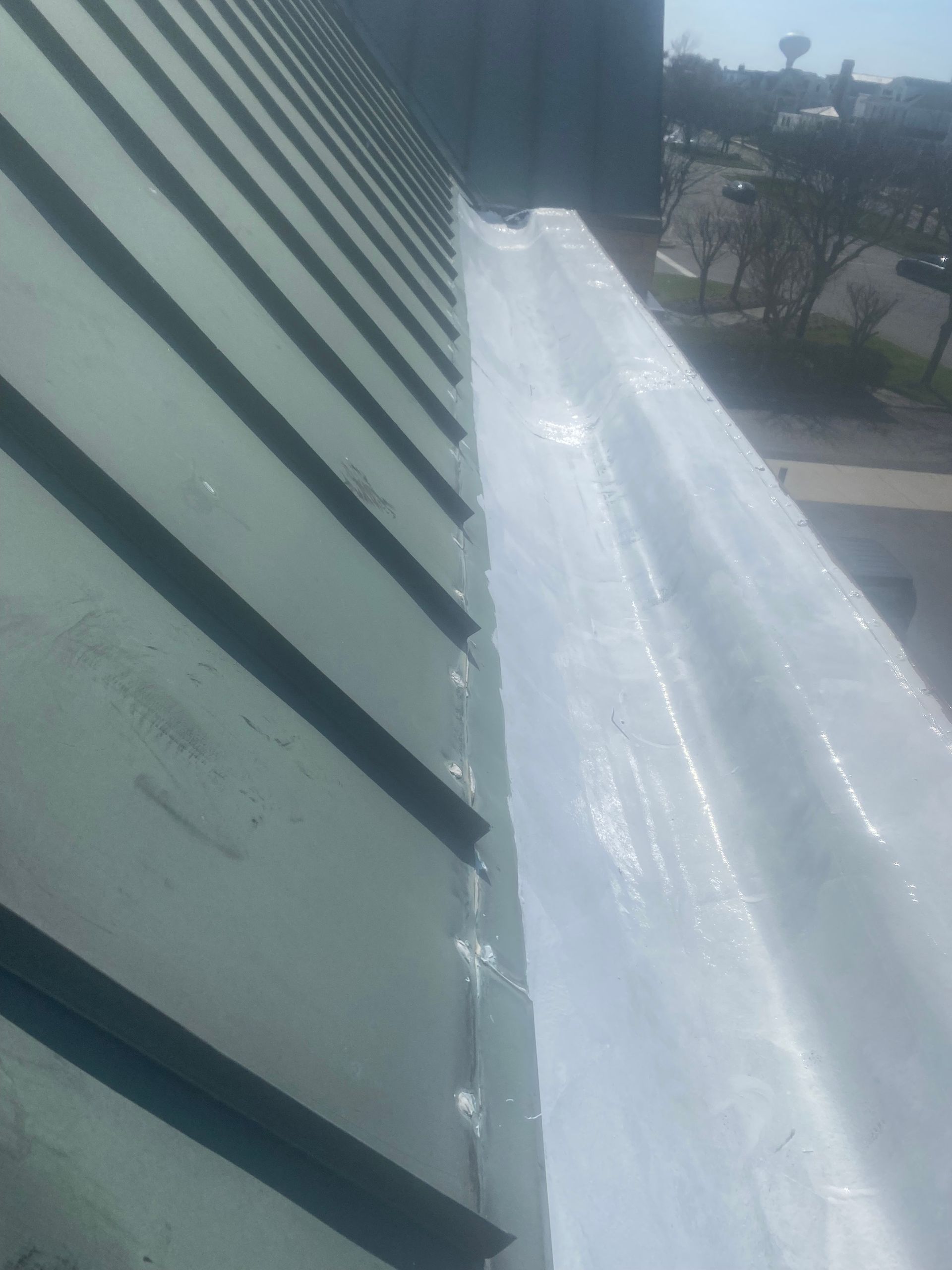 Tilted rooftop with green metal panels and a white tarp or membrane beside a road and trees below.