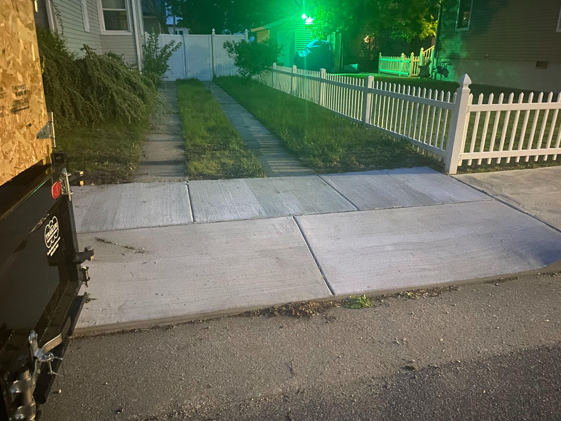 Nighttime driveway and sidewalk beside a white fence, lit by green streetlights.