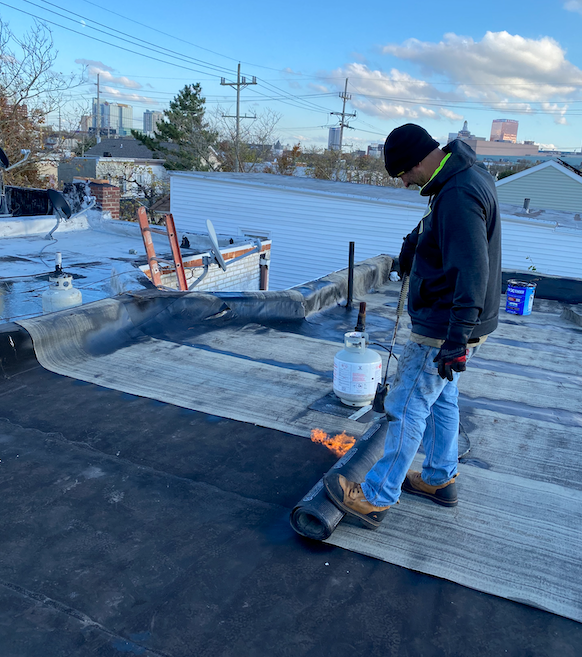 A man is standing on a roof with a roll of roofing material.