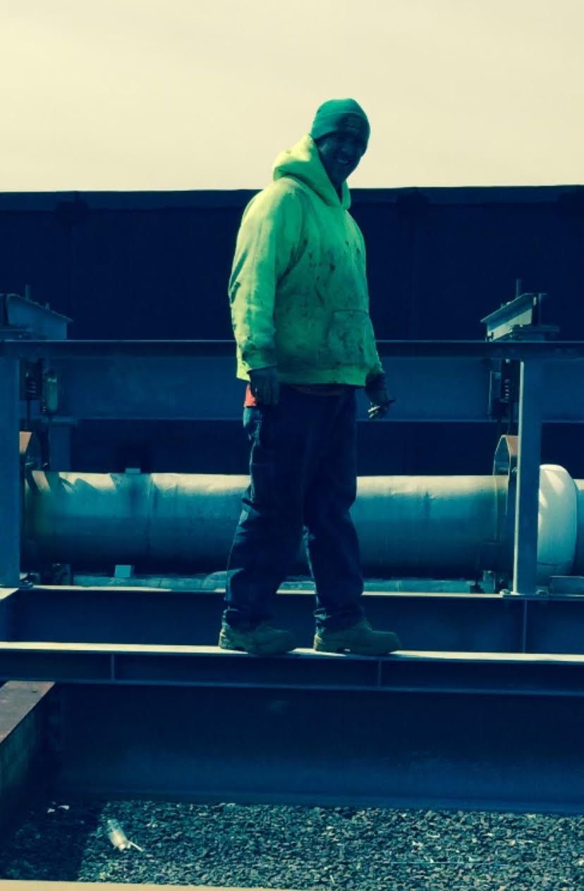Man in neon green hoodie stands on a platform above industrial equipment.
