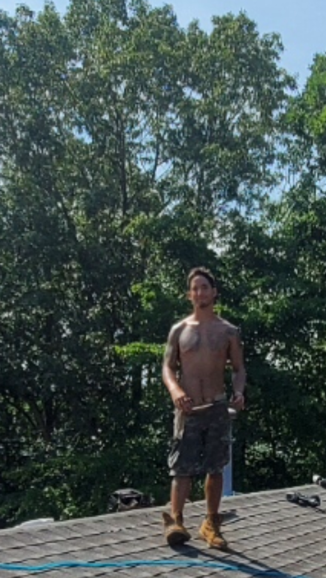 A shirtless man is standing on top of a roof holding a hammer.