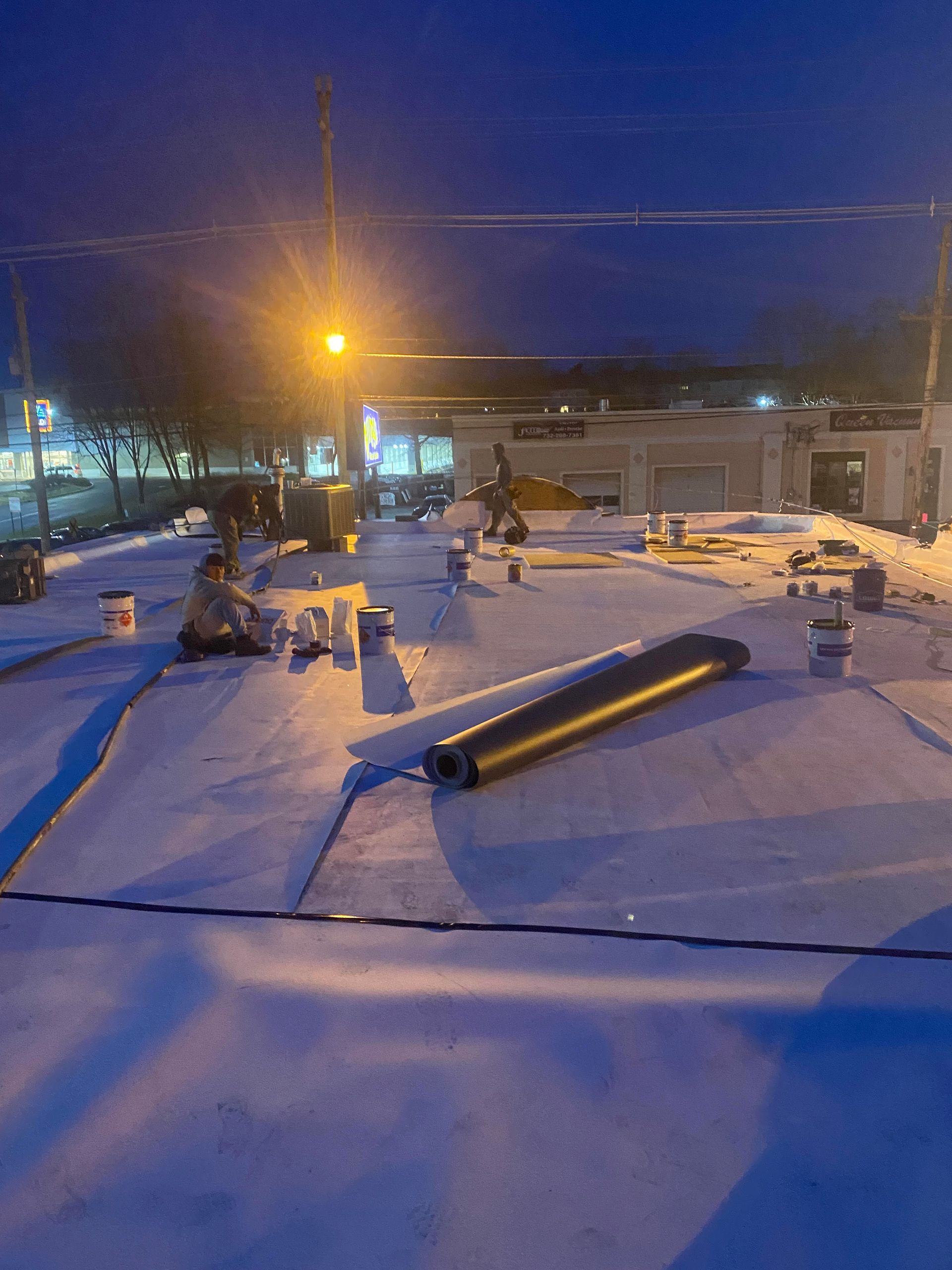 Workers on a commercial roof at dusk, rolling out roofing material, surrounded by buckets and equipment.