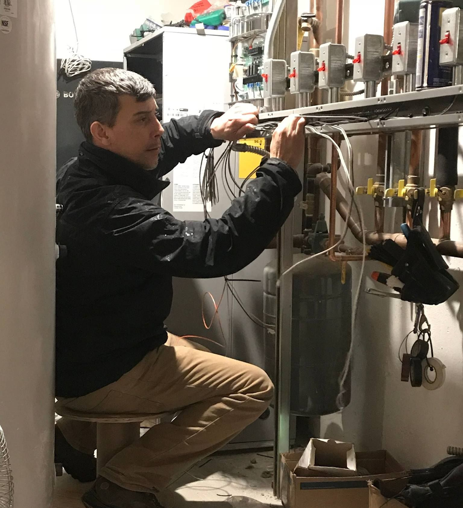Man in black jacket works on pipes in a utility room. He is kneeling on a stool.
