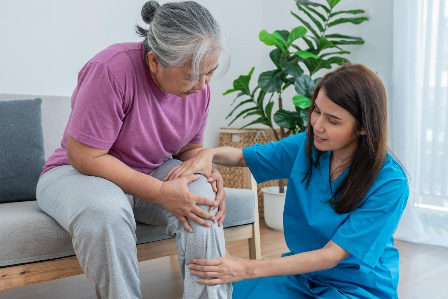 Caregiver in blue helping seated patient with knee support in a living room