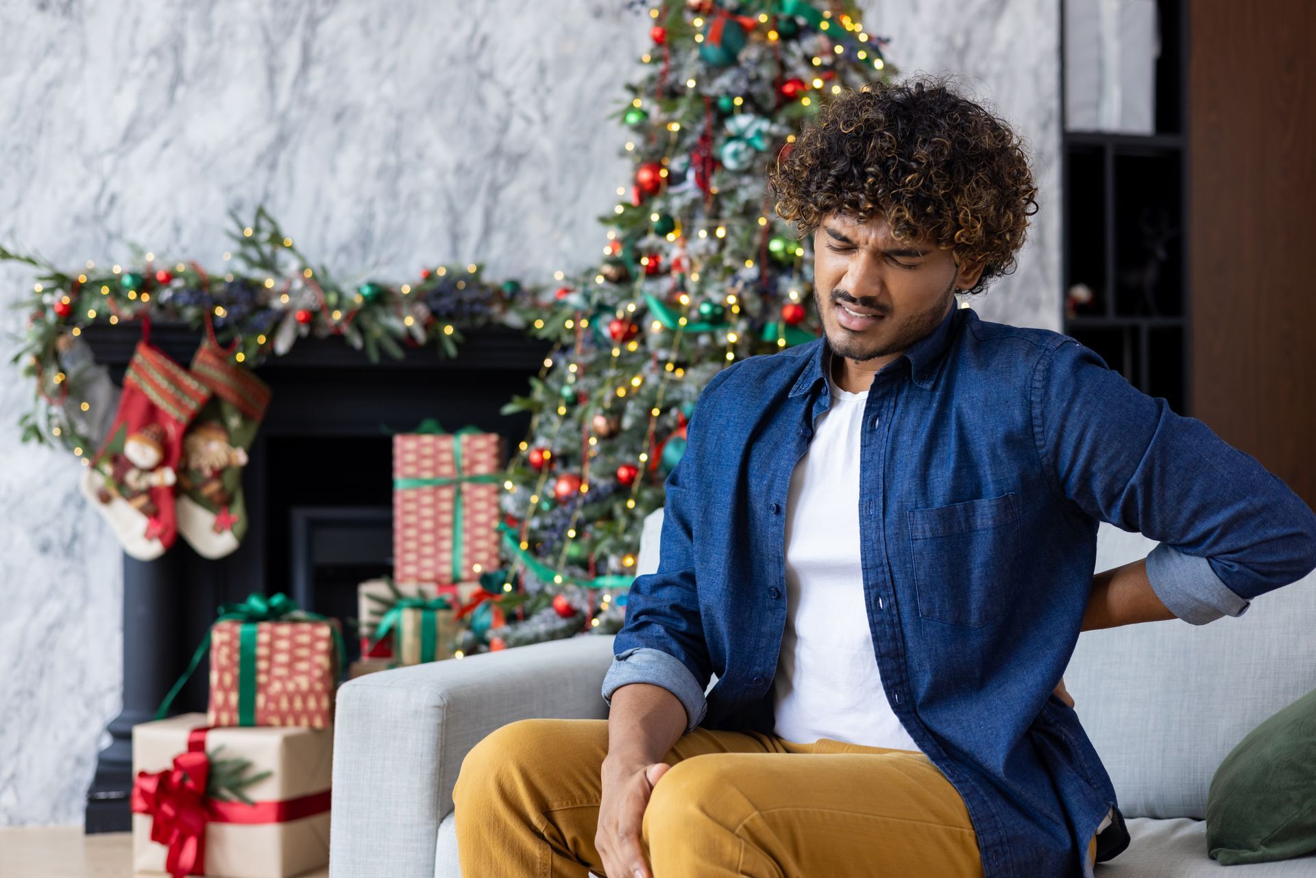 Man with back pain sits near decorated fireplace and Christmas tree.