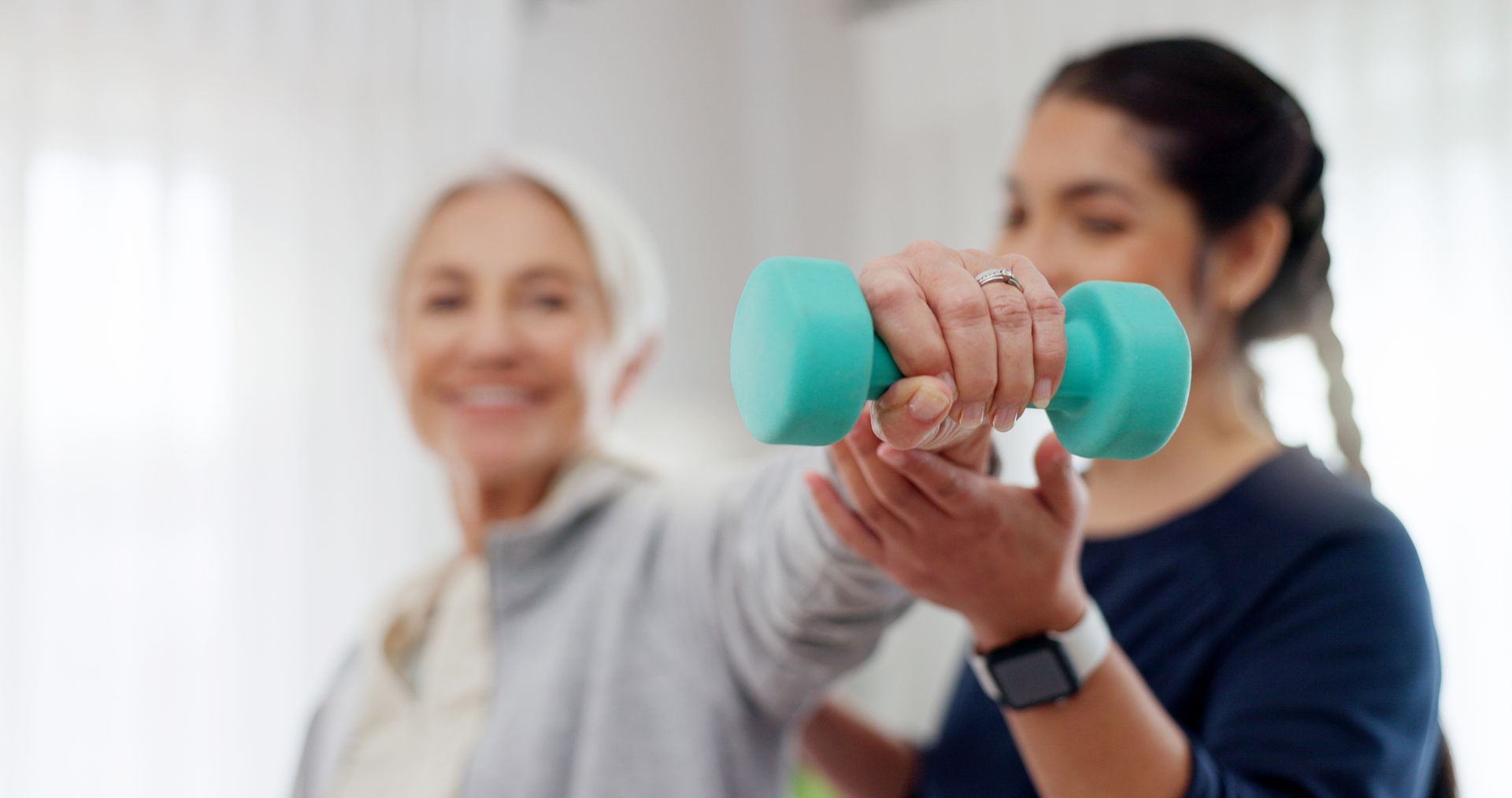 Senior woman exercises with dumbbell, guided by a young woman. Light blue dumbbell, smiling. Indoors.