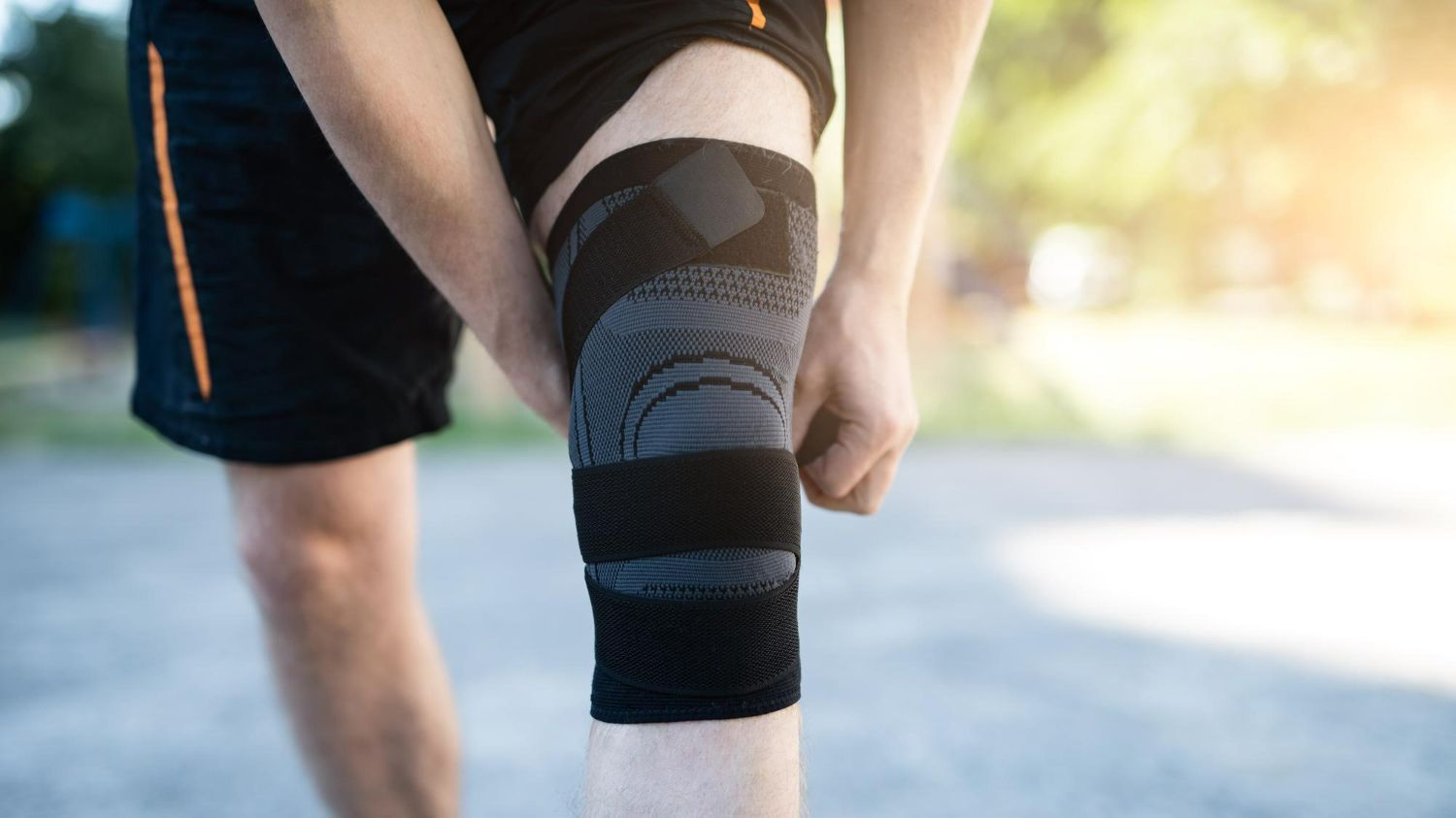 Person adjusting a black knee brace outdoors on a sunlit road