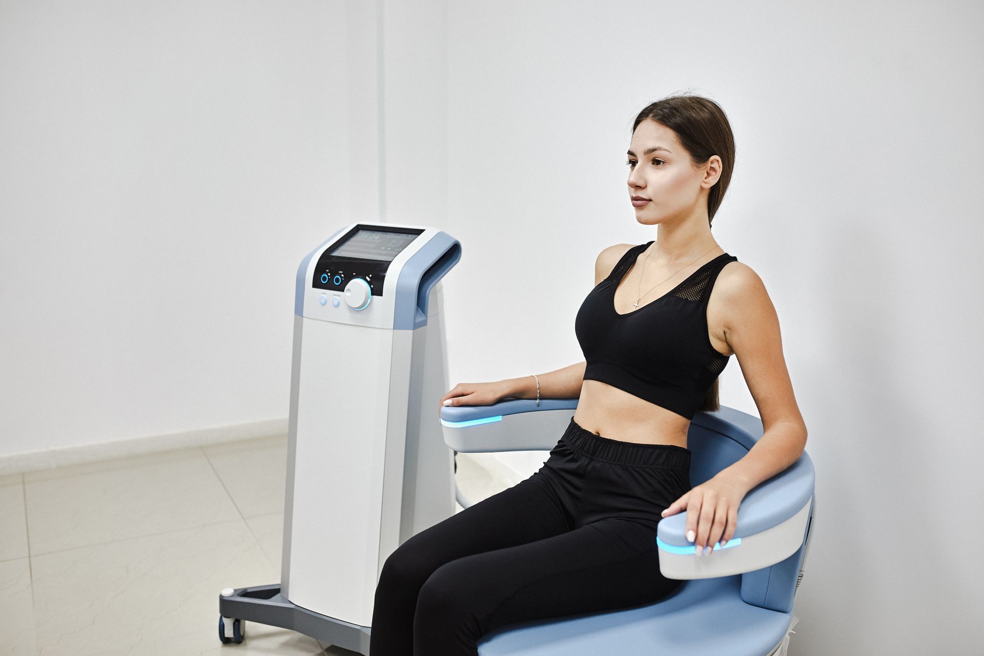 Woman seated in medical chair, next to a machine. She's wearing a black top and pants, focused. White walls.