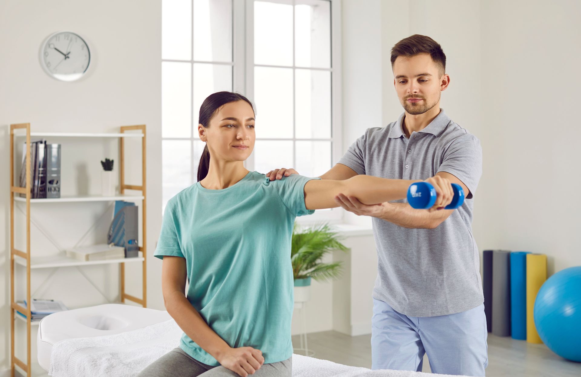 Therapist assists a patient lifting a dumbbell. The patient sits while extending their arm. The setting is a medical office.