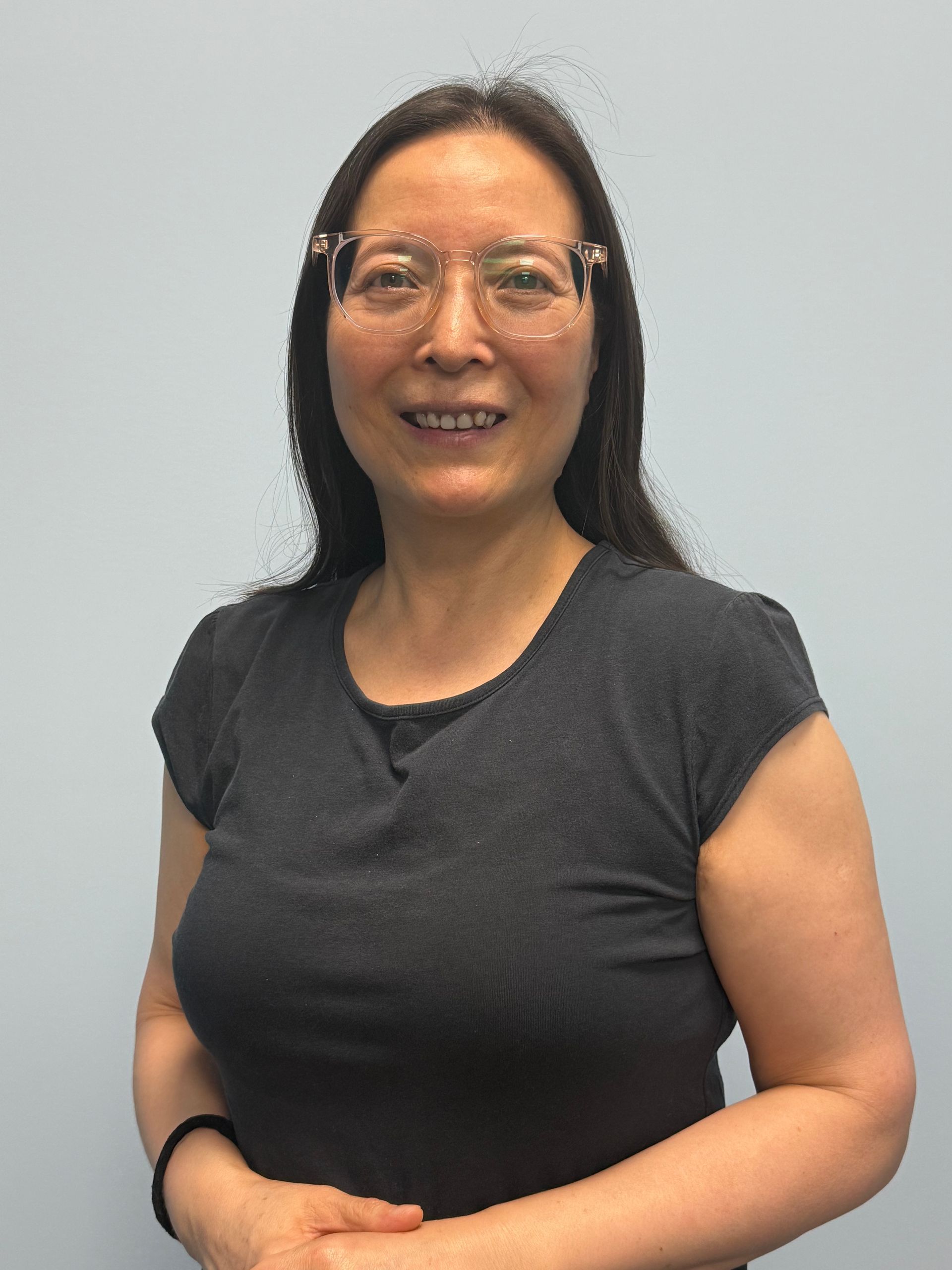 Woman with glasses smiles, wearing a dark gray shirt. Pale gray background.