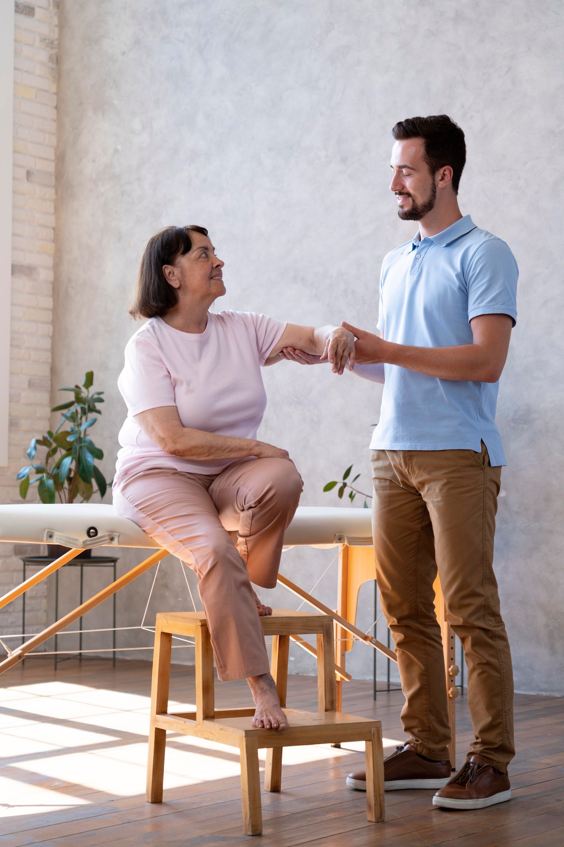 An elderly woman is sitting on a stool while a man holds her hand.